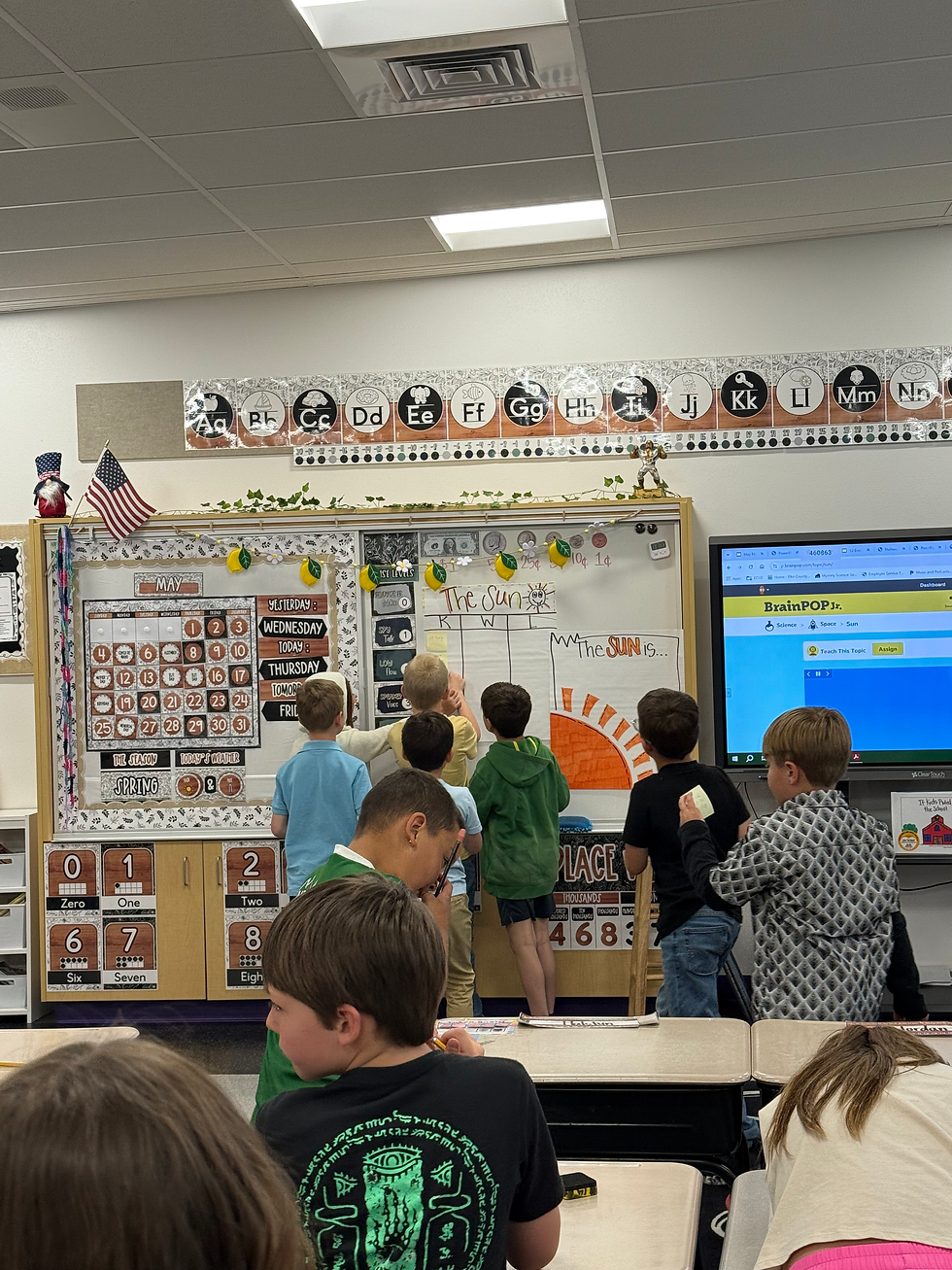 Children in a classroom face a board with sun-themed drawings. Bright colors and informational signs surround them, creating an energetic mood.