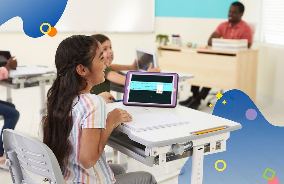 Girl in classroom, smiling at tablet on desk. Other children and teacher in background. Room is bright with colorful wall accents.