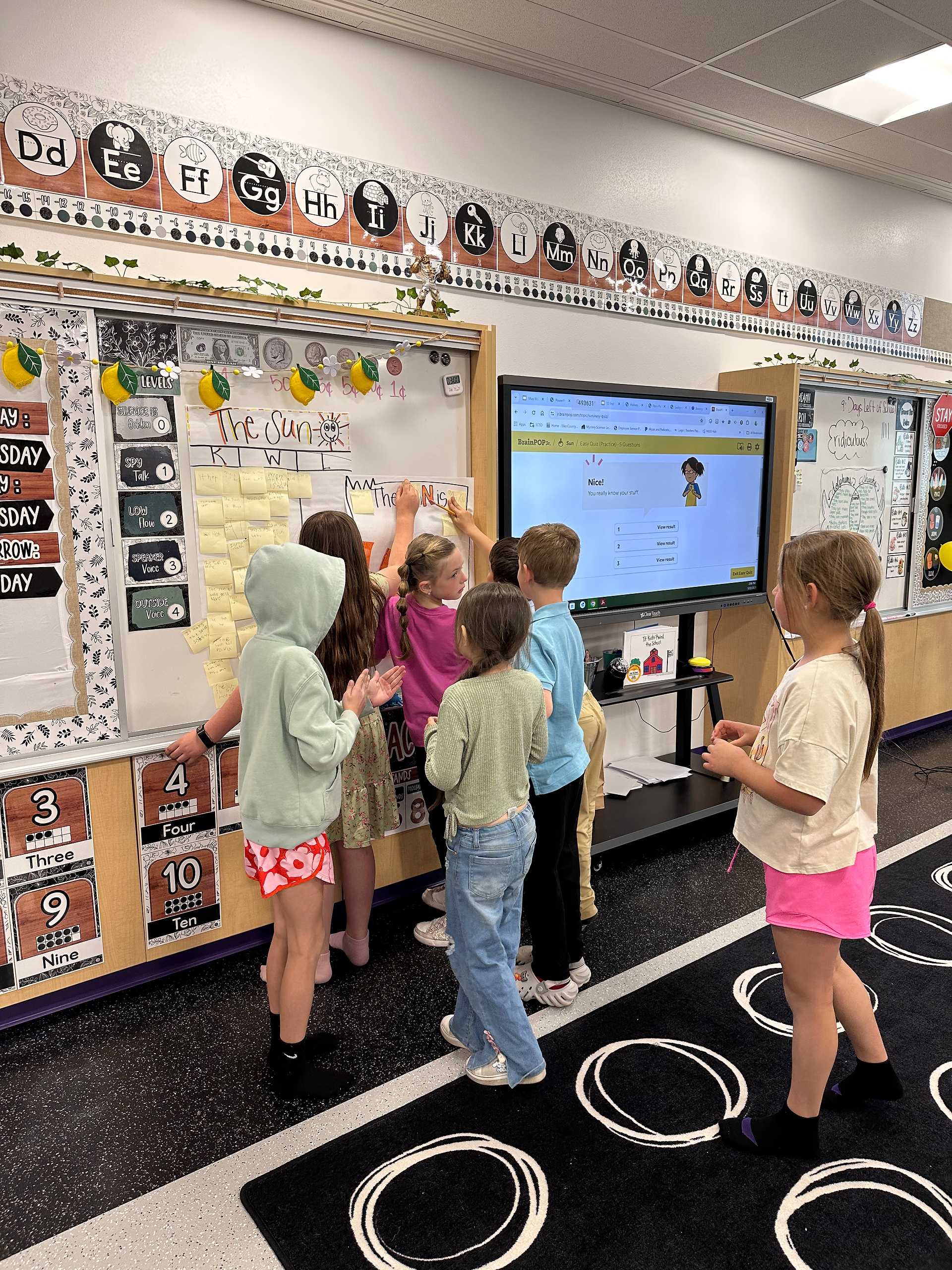 Young students come up to a white board in a classroom and place sticky notes on a KWL chart about the sun. BrainPOP Jr. quiz visible via Smartboard.